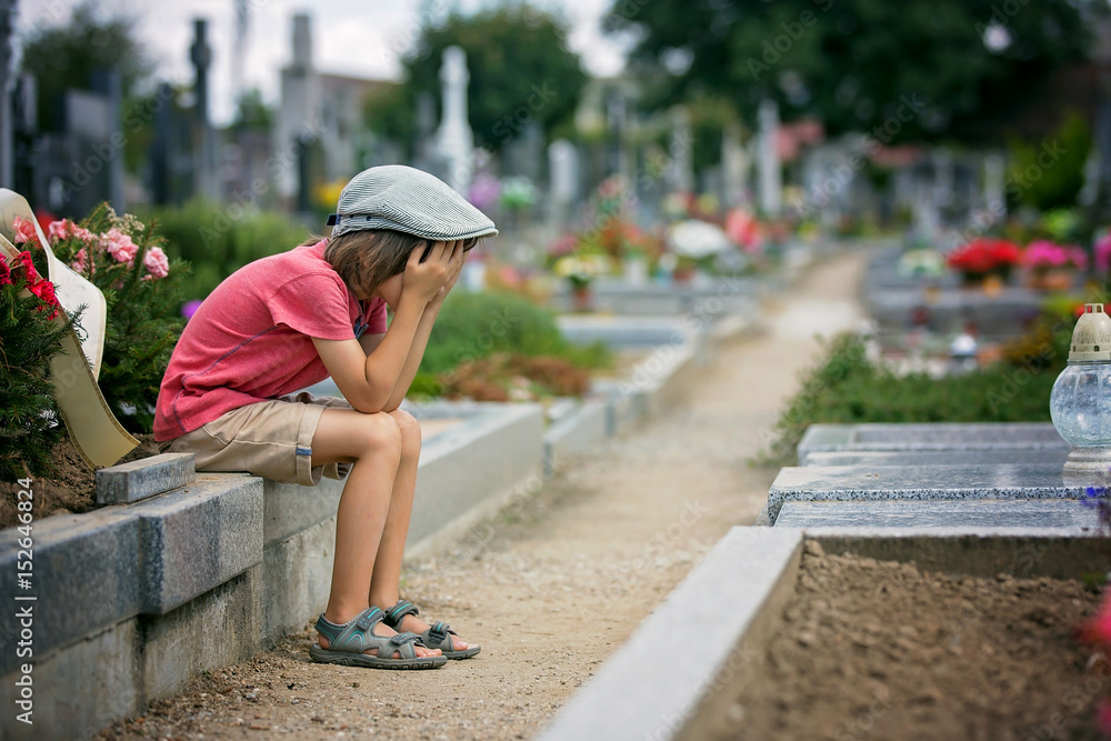 Sad little boy, sitting on a grave in a cemetery, feeling sad