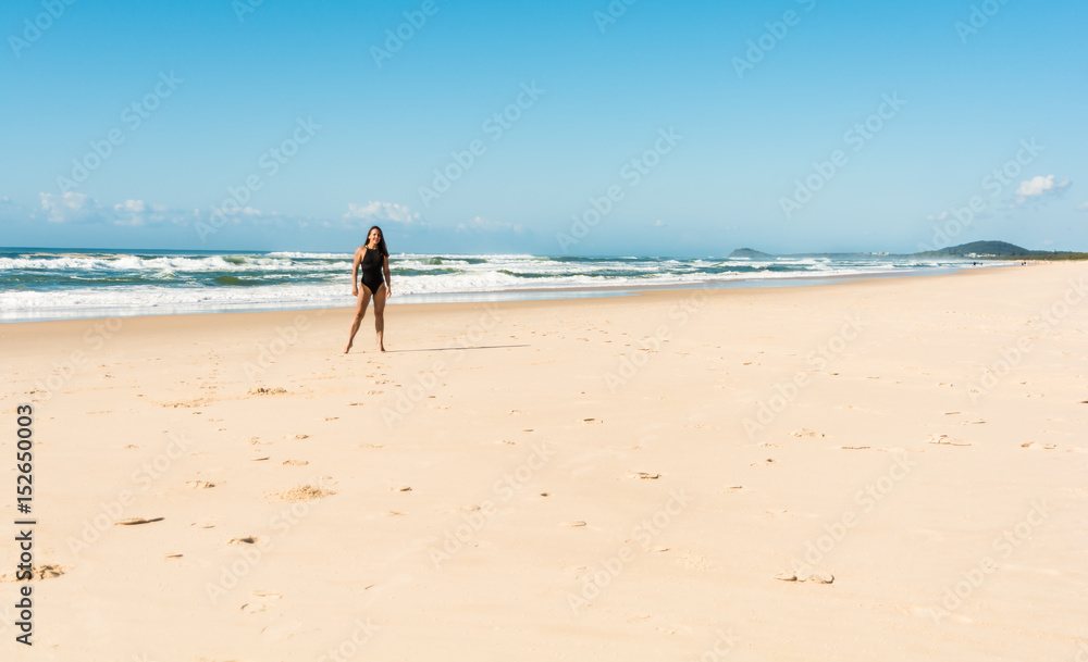The young woman girl with long dark hair wearing black swimsuit staying and posing on the beach with wonderful ocean background in sunny day in Byron Bay, Australia