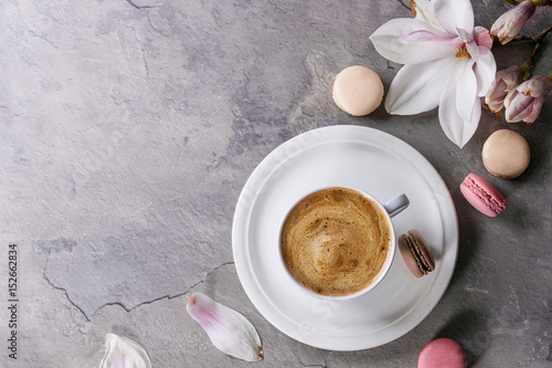 White cup of black coffee, served on white saucer with macaroons biscuits and magnolia flower blossom branch over gray texture background. Flat lay, space
