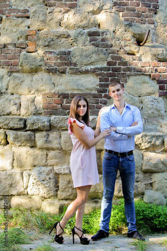 Woman and man posing at the camera. Stone and brick wall at the background. Girl dressed in pink dress and man in blue shirt and jeans.