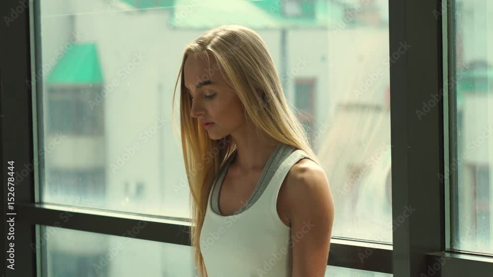 Attractive 20s blondy is engaged with dumbbells on the window background close-up. Attractive 20s blondy is engaged with dumbbells on the window background close-up.