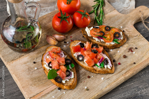 Italian bruschetta set. Varied sandwiches with cheese, tomatoes, fresh basil, olives, rosemary, spices. Bottle with olive oil,  on rustic wooden board over dark background,  top view