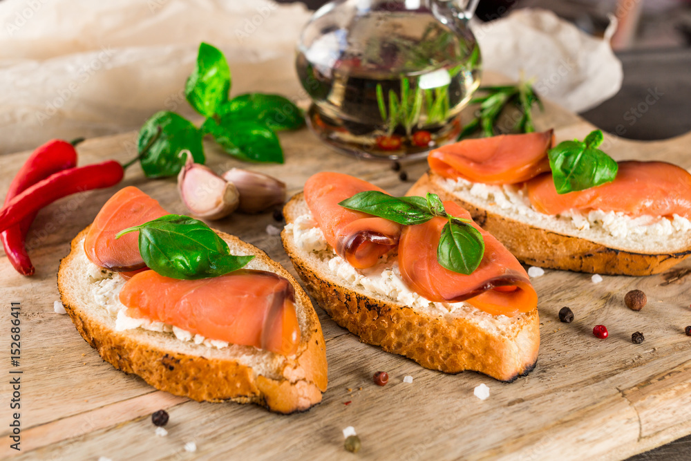 Italian bruschetta set. Sandwiches with cheese and red fish fresh basil, spices, chili peppers, rosemary, garlic. Bottle with olive oil,  on rustic wooden board over dark background,  top view