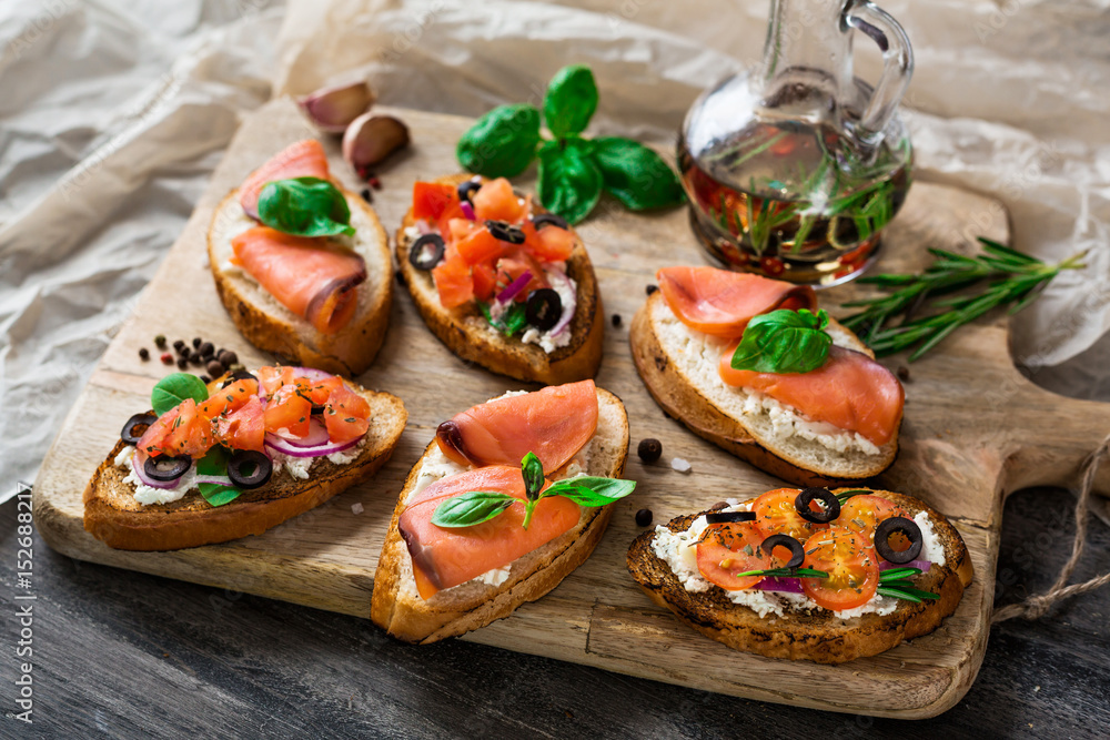 Italian bruschetta set. Varied sandwiches with cheese and red fish, roasted tomatoes, fresh basil, spices, rosemary, garlic, onion, chili pepper.  on rustic wooden board over dark background, top view
