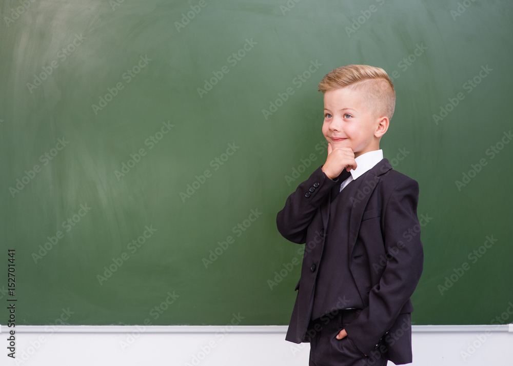 Thoughtful boy in a suit standing near empty green chalkboard Thoughtful boy in a suit standing near empty green chalkboard