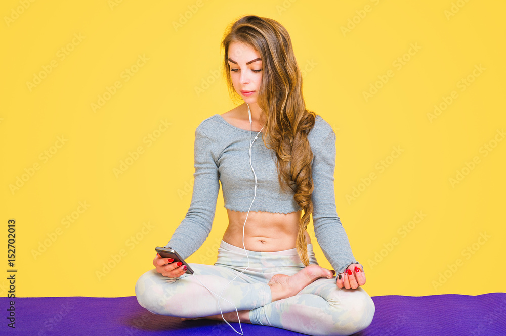 Beautiful young yogini woman sitting down in lotus pose on purple mat and listening to relaxing music from her phone Beautiful young yogini woman sitting down in lotus pose on purple mat and listening to relaxing music from her phone