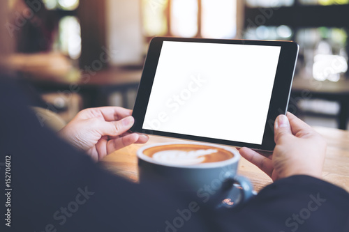 Mock up image of business woman's hands holding black tablet with white blank screen and coffee cup on wooden table in cafe