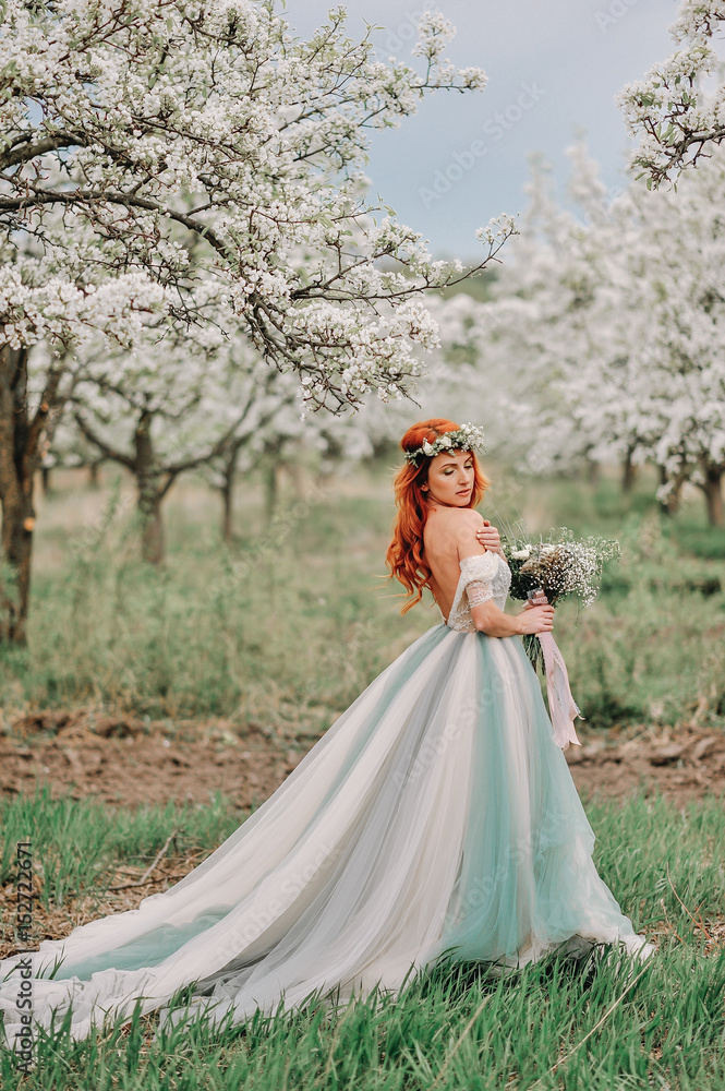 Young red-haired woman in a luxurious dress is standing in a blooming garden