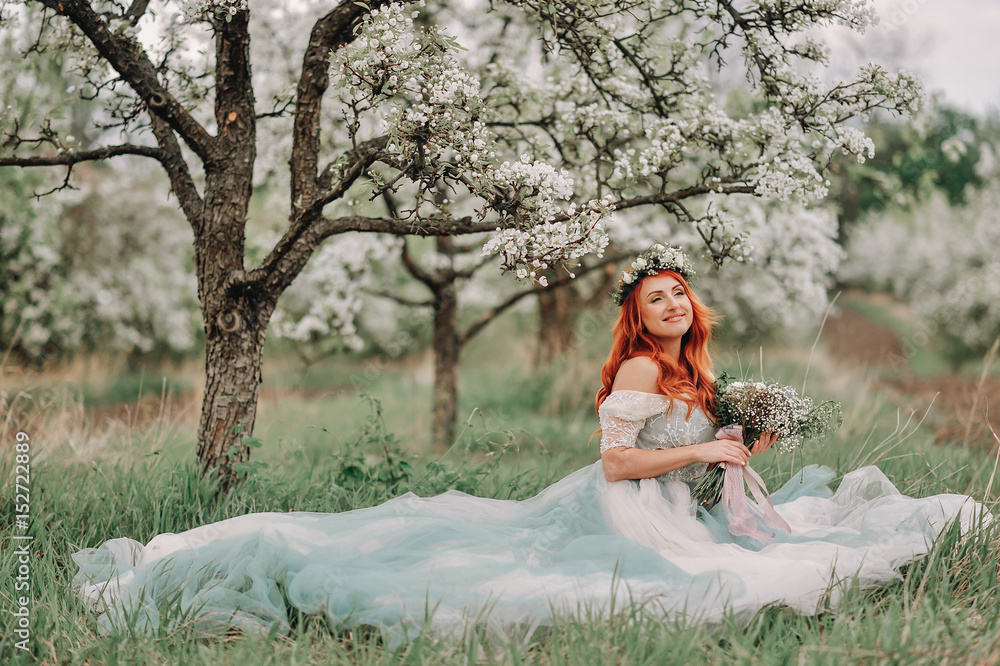 Young red-haired woman in a luxurious dress sits on the grass in a blooming garden