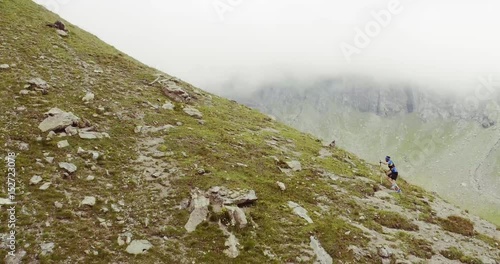 aerial flight above caucasian trail runner running to mountain through rocky climb.wild green nature outdoors in cloudy and foggy weather. 4k drone side wide passthrough video photo