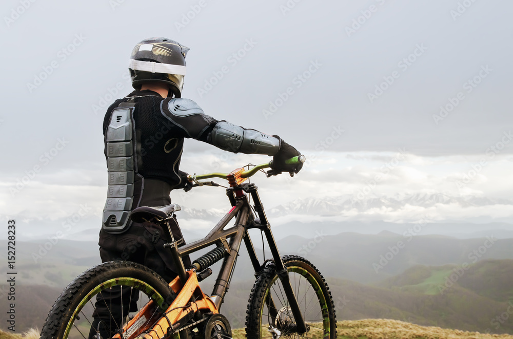 The rider in the full-face helmet and full protective equipment on the mtb bike stands on a rock against the background of a ridge and low clouds The rider in the full-face helmet and full protective equipment on the mtb bike stands on a rock against the background of a ridge and low clouds