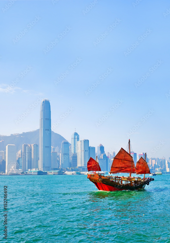 Naklejka premium Widok na panoramę Hongkongu z czerwoną chińską żaglówką przechodzącą na Port Wiktorii w słoneczny dzień.