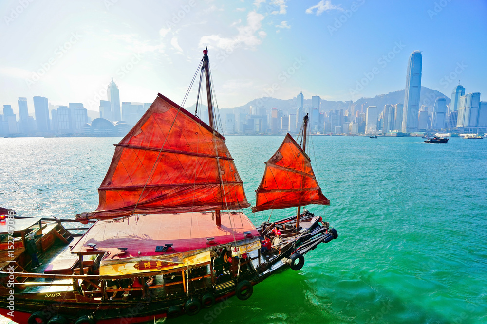 Fototapeta premium View of Hong Kong skyline with a red Chinese sailboat passing on the Victoria Harbor in a sunny day.