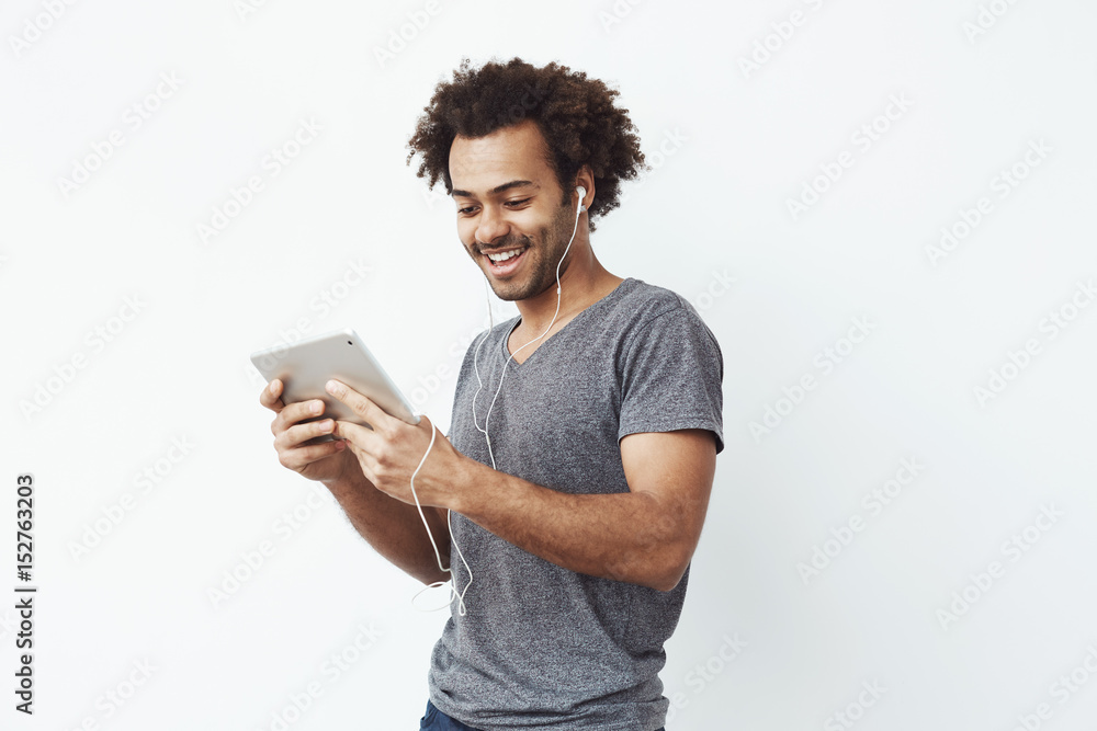 Portrait of cheerful happy african man in headphones laughing holding tablet talking or watching and enjoying a comedy show or browsing over white wall in wired headphones Portrait of cheerful happy african man in headphones laughing holding tablet talking or watching and enjoying a comedy show or browsing over white wall in wired headphones