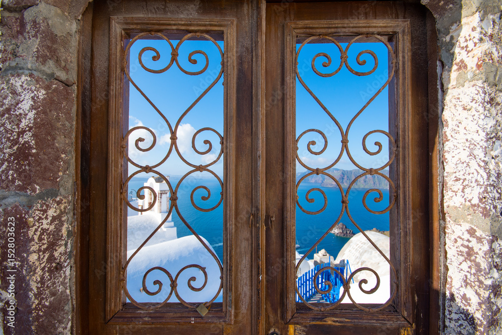 Oia town on Santorini island, Greece. Traditional and famous houses and churches with blue domes over the Caldera, Aegean sea. Frame through the window of a wooden door yard at the caldera. Oia town on Santorini island, Greece. Traditional and famous houses and churches with blue domes over the Caldera, Aegean sea. Frame through the window of a wooden door yard at the caldera.