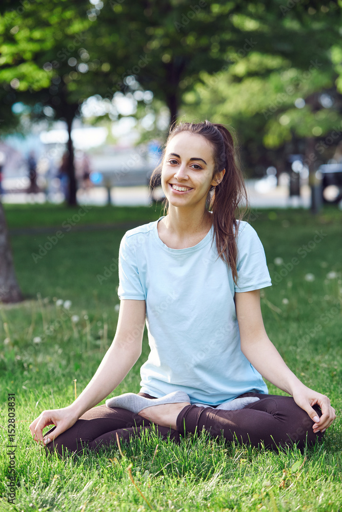 Portrait of young and sporty woman in sportswear doing yoga or Stretching exercises outside at the park on green field on cloudy day, Dnipro, Ukraine. She is sitting on the green meadow between trees Portrait of young and sporty woman in sportswear doing yoga or Stretching exercises outside at the park on green field on cloudy day, Dnipro, Ukraine. She is sitting on the green meadow between trees