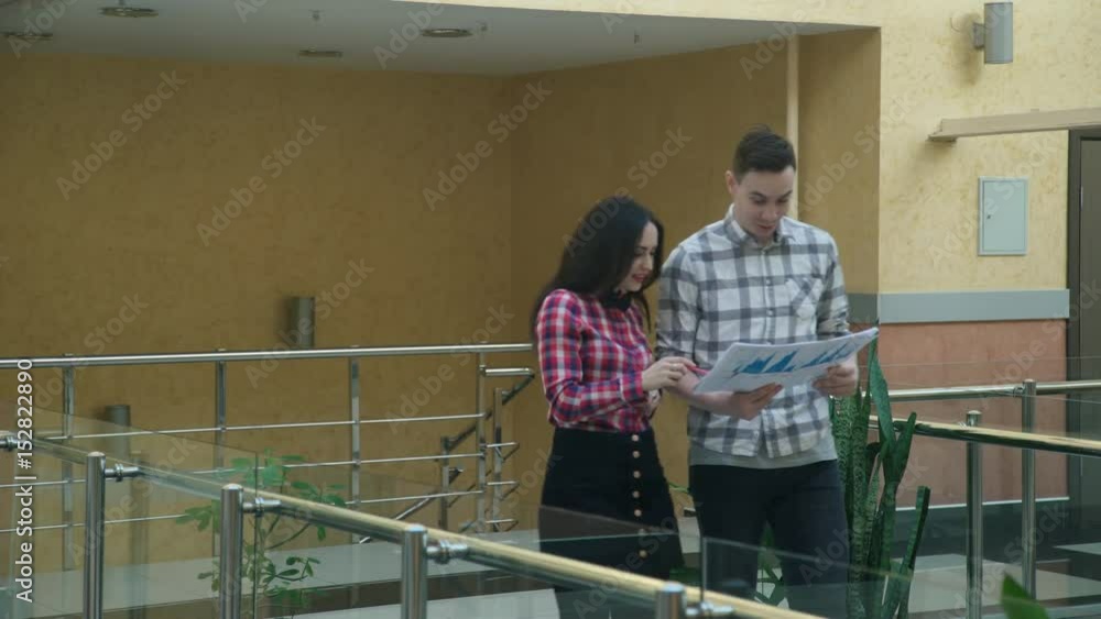 Woman man discussing topics, watching blueprints walking on hall. Cowokers communicate during break of working day in office. Concentrated brunette female with black hair in checkered red shirt, skirt Woman man discussing topics, watching blueprints walking on hall. Cowokers communicate during break of working day in office. Concentrated brunette female with black hair in checkered red shirt, skirt