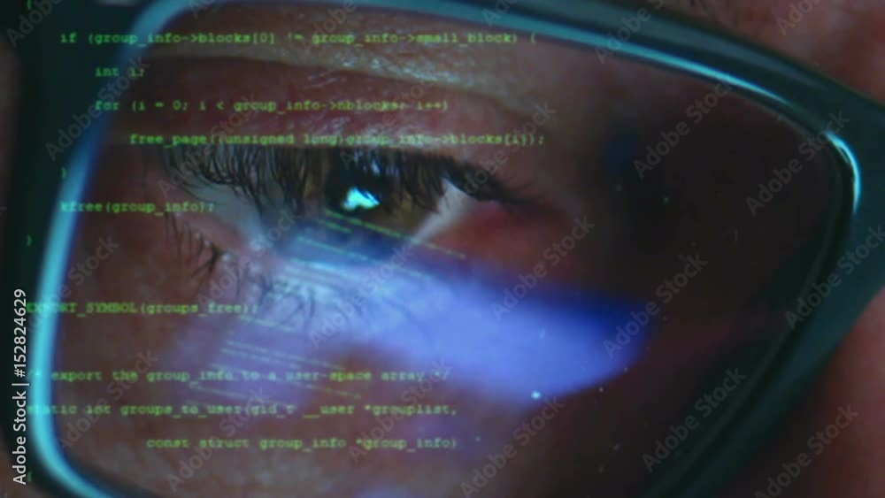 Male hacker working on a computer while green code characters reflect on his face in a dark office room