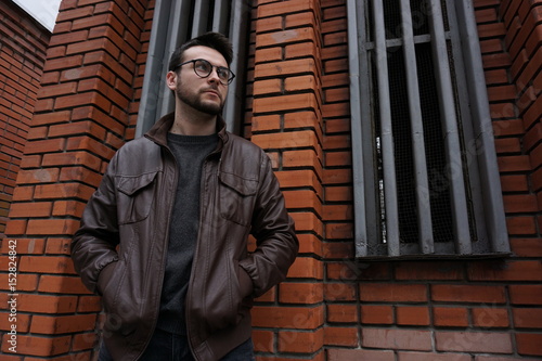 Handsome man with glasses. In a leather jacket. Brick background. A man looks in the side.
