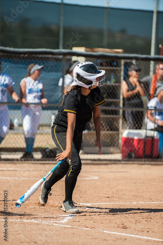 Teenage batter in black softball uniform dropping her bat after contact.  