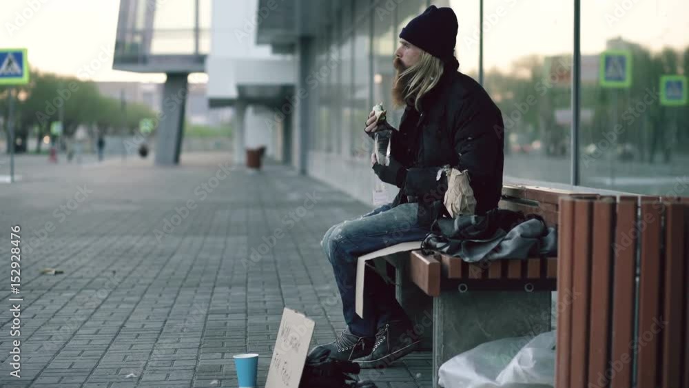 Homeless young man eating sandwich and drinking alcohol from paper bag on bench at city street in evening Homeless young man eating sandwich and drinking alcohol from paper bag on bench at city street in evening