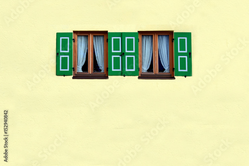 Yellow wall with two windows with green shutters open. Empty space below.
