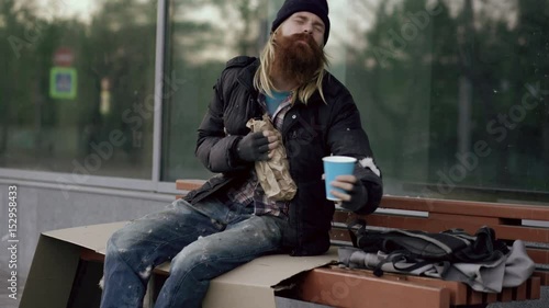 Very drunk homeless man talking to people walking near him and beg for money and show fuck you sign while sitting on bench at the sidewalk