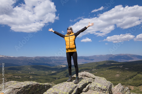 sport hiking or trekking woman with yellow jacket, standing on rock peak, with arms up and happy face, behind Lozoya Valley and Guadarrama Park, in Madrid, Spain 