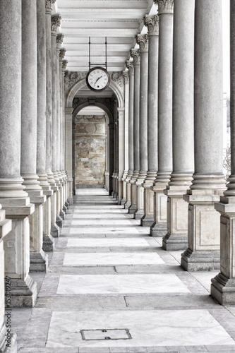 Fotografering Colonnade of tall columns with clock by the ceiling
