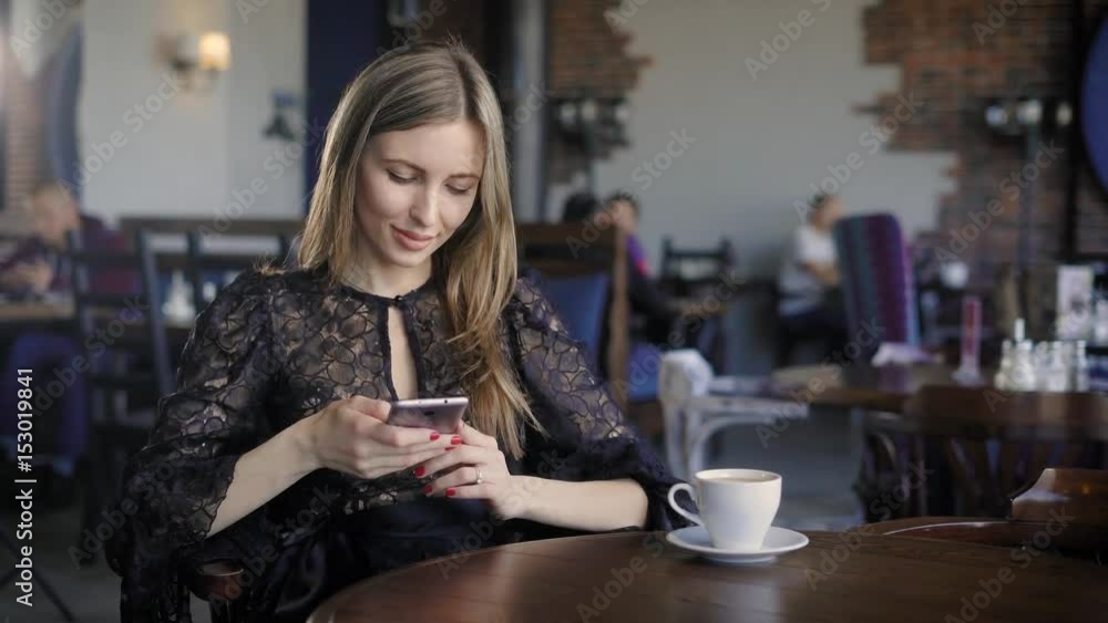Smiling female using phone. Young elegant girl sitting at table in cafe and using smartphone. Smiling female using phone. Young elegant girl sitting at table in cafe and using smartphone.