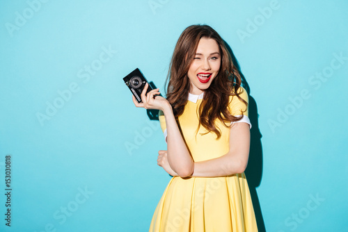Playful young woman in dress holding retro camera and winking
