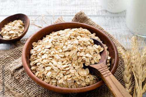Rolled oats or oat flakes in bowl with wooden spoons, golden wheat ears and bottle of milk on background. Healthy lifestyle, healthy eating concept