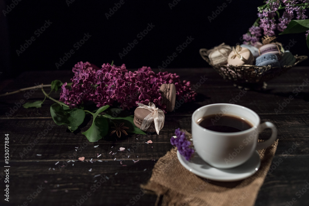 Cup of coffee on linen napkin and several macarons in braid vase with branch of lilac.