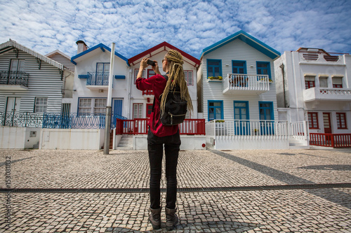 Traveller girl with blonde dreadlocks is takes photo on a smartphone striped houses in Costa Nova, Portugal