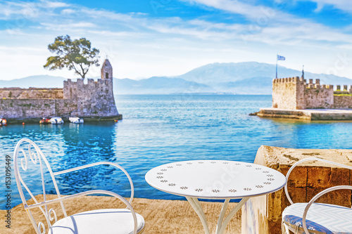 Table and chairs on coast of Nafpaktos old port bay in Greece, nearby Patras city. Incredible tourist landmark.