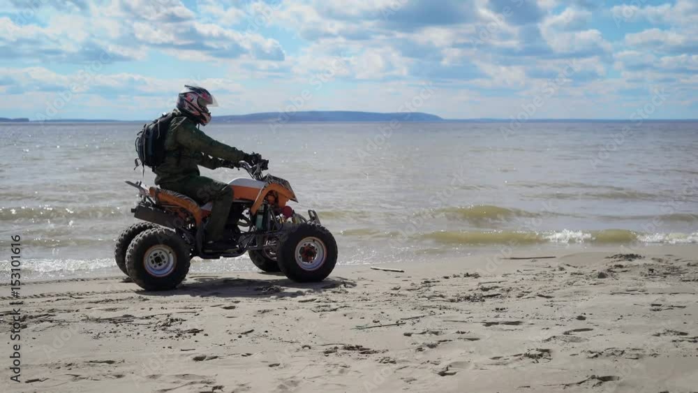 Man dressed in protective equipment and helmet riding ATV and driving along the seashore with high speed. Fan of extreme tourism with backpack behind rushing on his quad bike on the beach. Man dressed in protective equipment and helmet riding ATV and driving along the seashore with high speed. Fan of extreme tourism with backpack behind rushing on his quad bike on the beach.