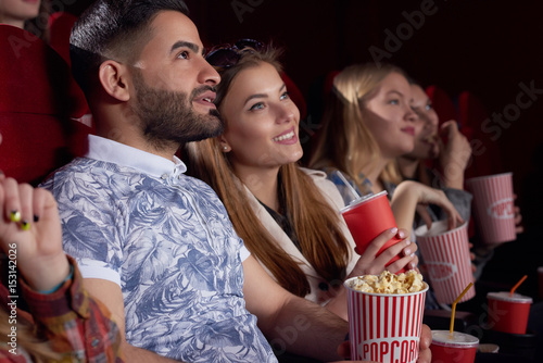 Side view of beautiful couple of caucasian girlfriend and arabian boyfriend watching interesting film at cinema hall, eating popcorn and drinking cola. Happy friends thinking about movie and smiling.
