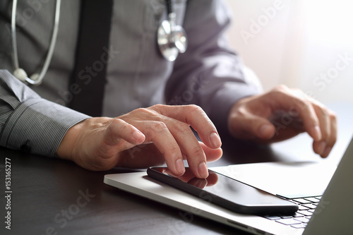 close up of smart medical doctor working with mobile phone and laptop computer and stethoscope on dark wooden desk