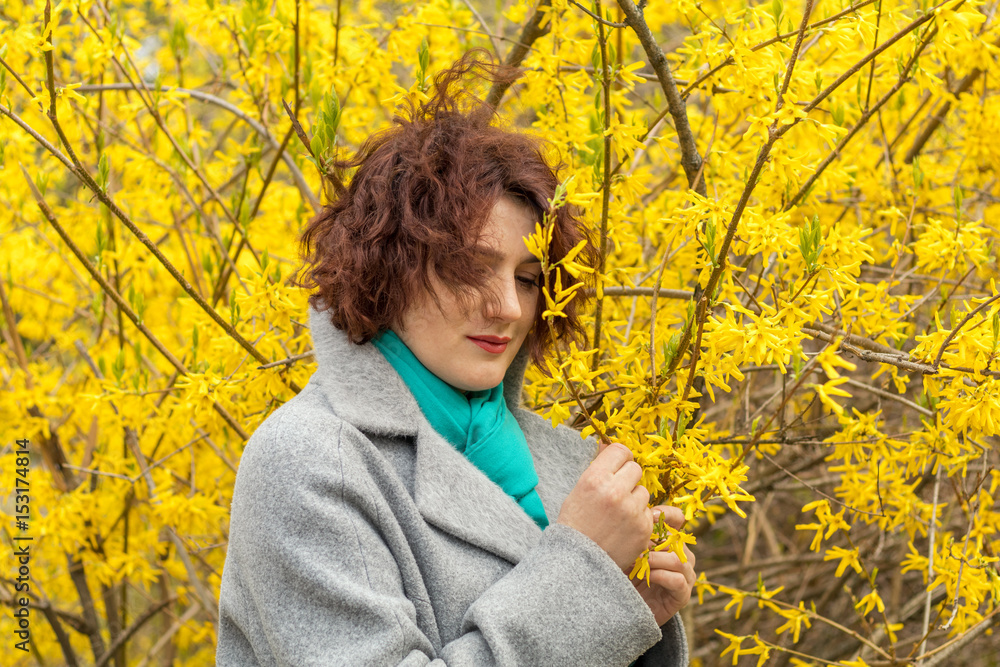 Pretty young red head woman with forsythia blossoms in spring park. Yellow blossoms background.