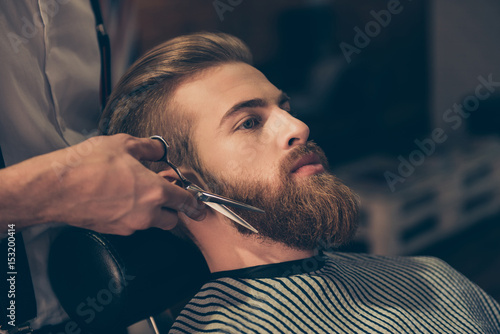 Close up of a hairdresser`s work for an attractive young man at the barber shop. He is doing styling of a red beard with scissors