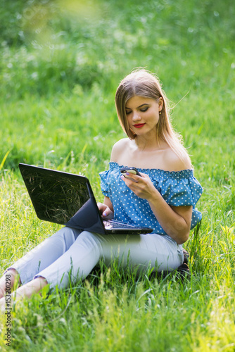 Young attractive woman with a laptop sitting on the grass and speaks by phone. © dmshpak