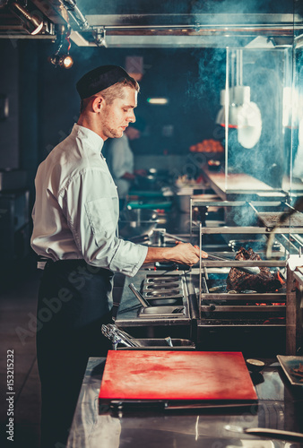 Food concept. Young handsome chef in white uniform monitors the degree of roasting and turns meat with the forceps in interior of restaurant kitchen. Preparing traditional beef steak on barbecue oven.