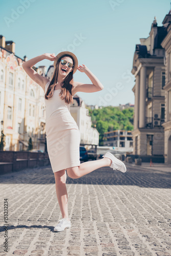 Cheerful dreamy girl jumping, posing for photo on vacation. She is so carefree, wearing hat, sunglasses, pure light dress