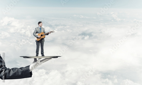 Businessman on metal tray playing acoustic guitar against blue sky background