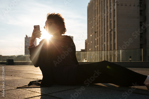 Curly african girl sitting outdoors on the roof taking mobile photos with sunset backlight over cityscape background