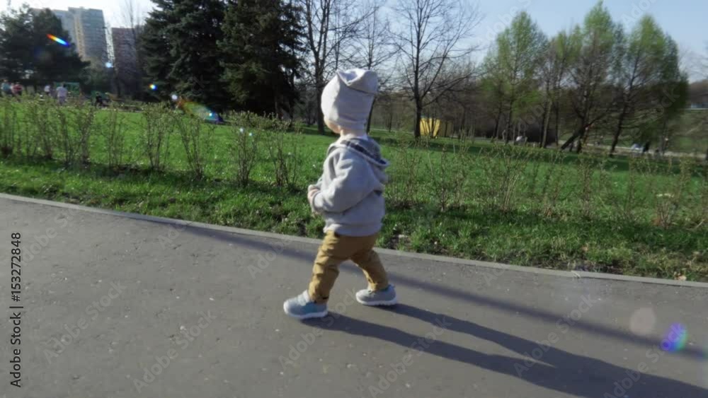 Little cute boy with confident rapid steps walks along the road in the park in summer at sunlight. In the background there are green lawns and trees. Beautiful summer day in the park Little cute boy with confident rapid steps walks along the road in the park in summer at sunlight. In the background there are green lawns and trees. Beautiful summer day in the park
