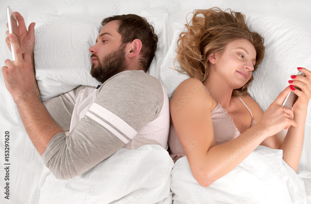 Young couple in bed looking phone and ignoring each other while lying together Young couple in bed looking phone and ignoring each other while lying together