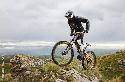 Ryder in full protective equipment on the mtb bike climbs on a rock against the backdrop of a mountain range and low clouds