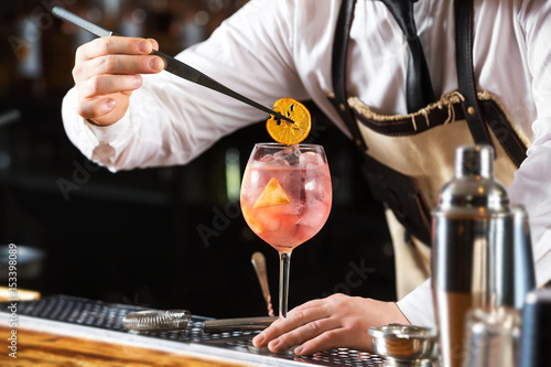 Elegant barman is making pink cocktail holding orange chips at bar counter background. photo