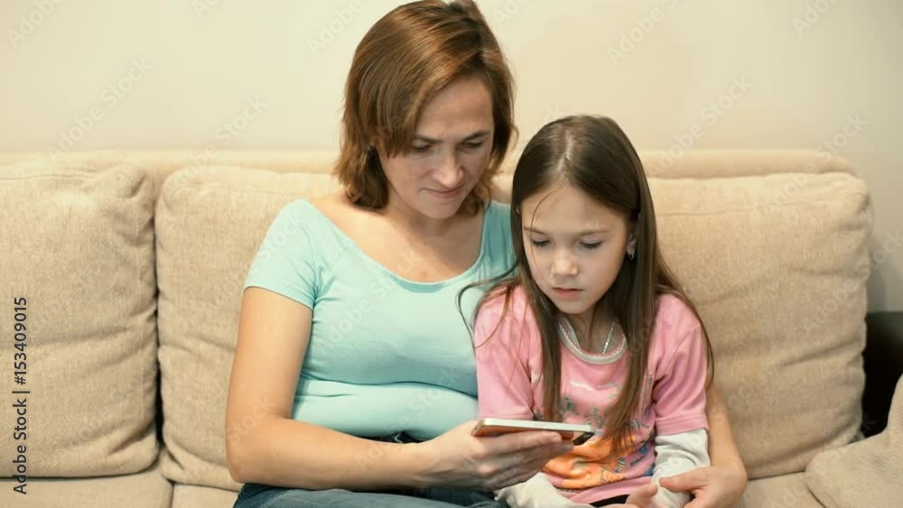 Mom and daughter sitting on sofa watching video on smartphone Mom and daughter sitting on sofa watching video on smartphone